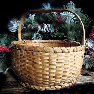 May include: A round, woven basket with a wooden handle. The basket is made of light brown woven strips. The background features evergreen branches, red berries, and pinecones, suggesting a rustic or holiday theme.