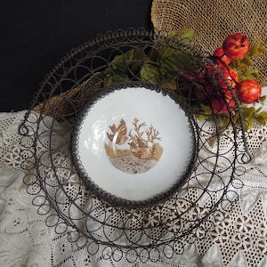 May include: A vintage white ceramic bowl with a brown bird and branch design, set inside a decorative, circular, dark metal wire basket. The bowl is surrounded by faux red apples and greenery, resting on a white lace tablecloth.