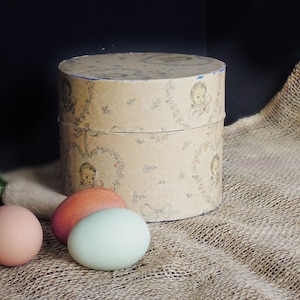 May include: A round cardboard box with a vintage design featuring illustrations of babies. The box is sitting on a burlap surface with three eggs: one brown, one orange, and one light blue.