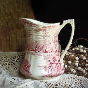 May include: A cream-colored ceramic pitcher featuring a detailed red transferware print of a rural landscape with houses, trees, and a body of water. The pitcher has a curved handle, a flared rim, and a wide base with a smooth, glossy finish.
