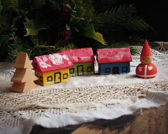 May include: A wooden Christmas scene featuring a small, light brown tree, three colorful houses with red, yellow, and green roofs, and a red and white gnome figurine. The houses have black door and window details. The scene is set on a lace cloth with greenery in the background.