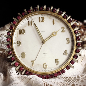 May include: Vintage round clock with a cream-colored face, gold hands, and gold numerals. The clock is surrounded by a gold bezel with red gemstones. The clock face has the text "SEVCA" and "7 JEWELS".
