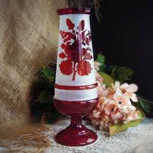 May include: A decorative glass vase with a white body and red accents. The vase features a floral design in red and has a flared top and a pedestal base. The vase is approximately 10 inches tall.
