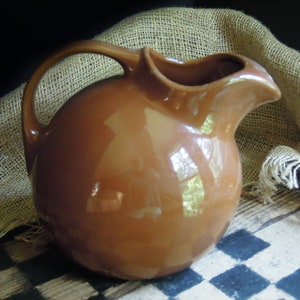 May include: A brown ceramic pitcher with a rounded body and a wide, curved spout. The pitcher is sitting on a checkered surface.