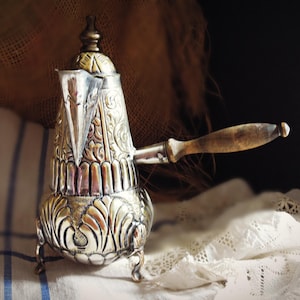 May include: Ornate silver-colored coffee pot with a wooden handle. The pot features intricate embossed floral designs and a decorative lid. The pot is resting on a white lace cloth, with a dark background.