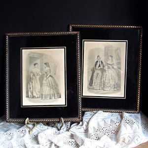 May include: Two framed vintage prints featuring women in elegant dresses. The prints are in black frames with gold trim, displayed on a white lace cloth. The artwork depicts women in long gowns, suggesting a historical or period theme.