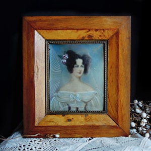 May include: A portrait painting of a person with dark hair styled in buns, wearing an off-the-shoulder white dress with a bow detail. The painting is encased in a rectangular wooden frame with a visible grain and a thin metallic inner border.