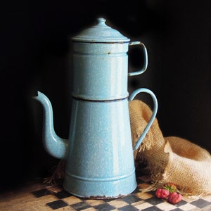 May include: A vintage, light blue enamelware coffee pot with a lid and handle. The pot has a classic design with a spout and a separate top section. It sits on a checkered surface with a burlap sack and a few strawberries in the background.