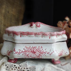 May include: A white ceramic trinket box with a lid, featuring a red floral design and trim. The box has a scalloped edge and sits on small feet. The overall aesthetic is vintage and decorative.