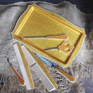 May include: A collection of vintage gold glitter hair styling tools, including combs, scissors, and a hook, arranged on a matching tray. The tools have white and gold glitter accents. The background is a burlap cloth.