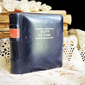 May include: A small, square, dark navy blue book with a glossy finish. The front cover features the text "BOOK OF TIME" in white, centered between two horizontal lines. The spine has a red rectangular label with gold text reading "BOOK OF TIME".