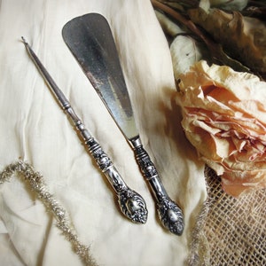 May include: Two silver-toned tools, a shoehorn and a crochet hook, are displayed on a white fabric background. The shoehorn has a decorative handle and the crochet hook has a long, slender handle.