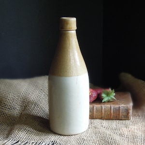 May include: A tall, antique stoneware bottle with a light beige top and a cream-colored body. The bottle is displayed on a burlap cloth with a vintage book and strawberries.