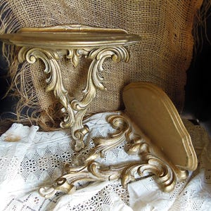 May include: Two ornate, gold-colored wall shelves with intricate scrollwork. One shelf is mounted, while the other rests on a white lace cloth. The shelves have a vintage, antique aesthetic, perfect for displaying small items.