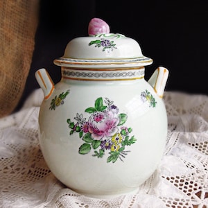 May include: A white ceramic sugar bowl with two handles and a matching lid featuring a pink floral finial. The body is decorated with hand-painted clusters of pink, yellow, and purple flowers with green leaves and a black geometric border pattern.