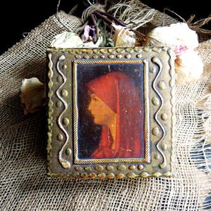 May include: A square decorative box featuring a central portrait of a person in profile wearing a red head covering. The box has a metallic gold finish with a raised border pattern of wavy lines and dots surrounding the central rectangular image.