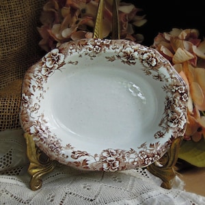 May include: An oval ceramic bowl featuring a white center and a decorative brown floral pattern along the scalloped rim. The bowl is supported by a gold-toned metal stand with ornate, curved legs, showcasing a detailed botanical design.