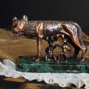 May include: Bronze sculpture of a she-wolf suckling two human infants, set on a green marble base. The wolf is detailed with textured fur and a fierce expression. A classic Roman art piece.