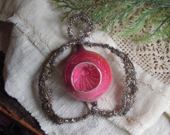 May include: A vintage Christmas ornament with a red and pink glass bulb, accented with silver tinsel. The ornament has a unique shape and is likely from a bygone era. The background includes a textured fabric and greenery.