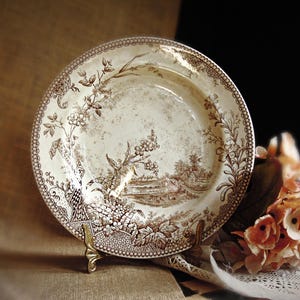 May include: A round ceramic plate featuring a brown transferware pattern on a cream background. The design depicts a detailed landscape with trees, foliage, and a small building, framed by a decorative border of repeating geometric shapes.