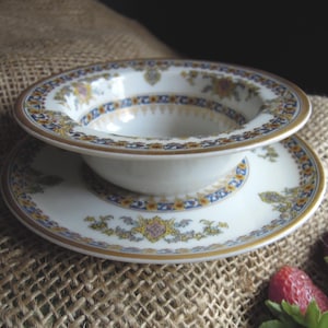 May include: A white porcelain bowl and saucer set with intricate gold, blue, and yellow floral patterns. The bowl sits atop the saucer, both displaying a detailed design. The set is on a textured, neutral-toned surface.