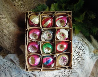 May include: A vintage box of Christmas ornaments. The ornaments are in various colors, including red, pink, green, and gold. Each ornament has a unique design, with stripes, swirls, and glitter. The box is made of cardboard and has individual compartments for each ornament.