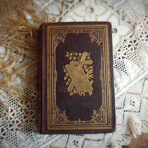 May include: Antique brown leather-bound book with gold embossed cross and floral design on the cover. The book is rectangular and has ornate gold detailing around the edges. The book is resting on a white lace tablecloth.