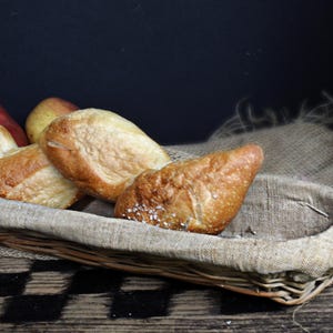 May include: A rustic wicker basket filled with assorted baked bread rolls and two pears. The bread has a golden-brown crust, with some rolls sprinkled with salt. The basket is lined with burlap, and the background is dark.