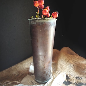 May include: A tall, textured, brown vase holds a small arrangement of red berries and green leaves. The vase has a ribbed design and a slightly flared top. The background is dark, and the vase sits on a burlap cloth.