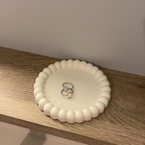 Pode incluir: Uma bandeja branca com borda ondulada contém dois anéis de prata. O prato é redondo e está sobre uma prateleira de madeira. Os anéis têm um design simples e elegante, ideal para guardar joias.