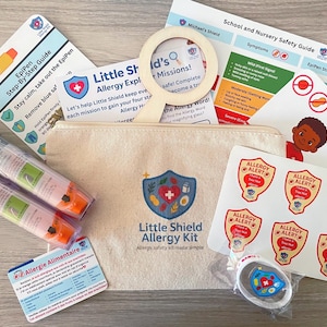 May include: A collection of items from the "Little Shield Allergy Kit", featuring EpiPens, educational guides, stickers, and a magnifying glass. The kit, designed for allergy management, includes a beige canvas pouch with a logo.