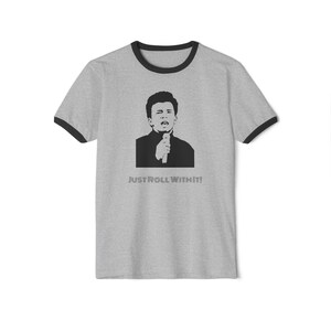 May include: A gray ringer t-shirt with black trim around the collar and sleeves. The shirt features a black and white graphic of a person holding a microphone, with the text "JUST ROLL WITH IT!" printed below the graphic.