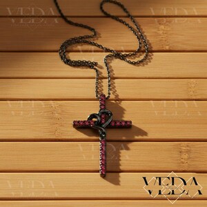 May include: A black metal cross pendant necklace with a heart-shaped design. The cross is adorned with small, red gemstones. The necklace hangs on a chain against a wooden background.