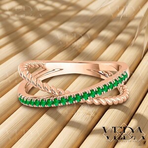 May include: Rose gold ring with a twisted band design. The ring features a row of small, vibrant green gemstones set along the top band. The ring's design is a modern take on a classic style.