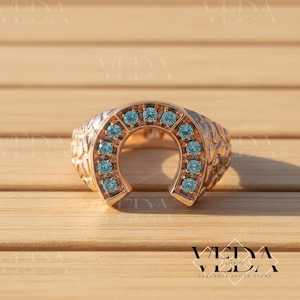 Könnte beinhalten: Ein roségoldener Hufeisenring mit hellblauen Edelsteinen. Der Ring hat ein strukturiertes Band und liegt auf einem Holzgrund. Der Text "VEDYA" und "ELEGANCE SET IN STONE" sind sichtbar.