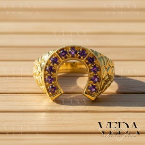 May include: A gold-colored horseshoe ring with a textured band and a row of purple gemstones. The ring is set against a wooden background, with the brand name "VEDA" visible in the lower right corner.