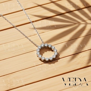 May include: A silver necklace with a circular pendant. The pendant is adorned with multiple clear gemstones set in a circle. The necklace is displayed on a wooden surface with a leaf shadow.