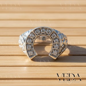 Puede incluir: Un anillo de herradura plateado con una fila de piedras preciosas transparentes en el centro. El anillo tiene un diseño texturizado en los lados y se muestra sobre una superficie de madera. El anillo es un símbolo de buena suerte.