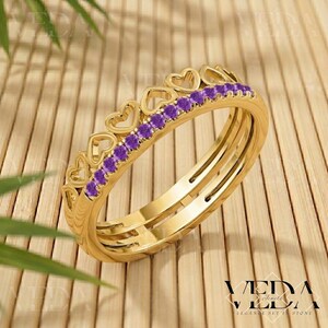 Könnte beinhalten: Ein goldener Ring mit einer Reihe von lila Edelsteinen und herzförmigen Aussparungen. Der Ring hat ein dreifaches Banddesign und ist vor einem Holzgrund platziert. Der Ring ist ein Schmuckstück.