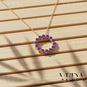 May include: A gold-colored necklace featuring a circular pendant adorned with purple gemstones. The pendant is suspended from a delicate chain. The necklace is displayed on a light brown wooden surface.