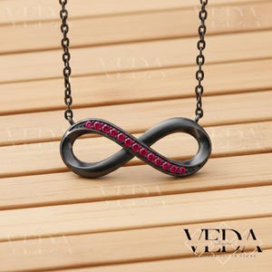 May include: A black infinity symbol pendant necklace with a chain. The infinity symbol is adorned with small, red gemstones. The necklace is displayed on a wooden surface. The necklace is a piece of jewelry.