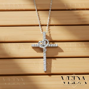 May include: A silver-toned pendant necklace featuring a cross design with a heart-shaped embellishment. The cross and heart are adorned with sparkling, clear gemstones. The necklace hangs on a delicate chain, set against a wooden background.