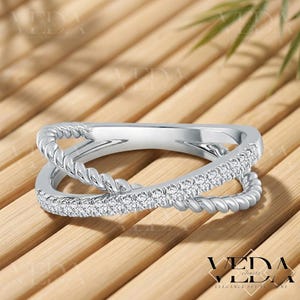 May include: A silver-toned ring with a crossover design. One band is adorned with small, sparkling stones, while the other features a twisted rope texture. The ring is set against a wooden background.