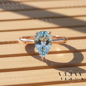 May include: A rose gold ring featuring a pear-shaped, light blue gemstone. The gemstone is the focal point, set on a delicate band. The ring is displayed on a wooden surface, showcasing its elegant design.