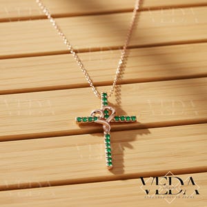 May include: Rose gold cross pendant necklace with emerald green gemstones. The cross features a heart-shaped design at the top and is set on a delicate chain. The necklace is displayed on a wooden surface.