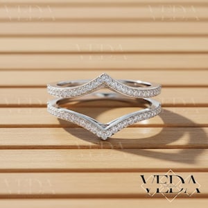 Puede incluir: Dos anillos plateados en forma de V, incrustados con pequeñas piedras preciosas transparentes. Los anillos están colocados sobre una superficie de madera marrón claro. Los anillos están diseñados para usarse juntos.