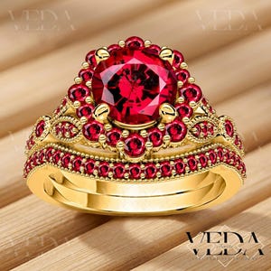 Op de afbeelding: Een goudkleurige ringenset met een grote, ronde, gefacetteerde rode edelsteen in het midden, omringd door kleinere rode edelstenen. De band is ook versierd met rode edelstenen. De ringenset wordt gepresenteerd op een houten oppervlak.