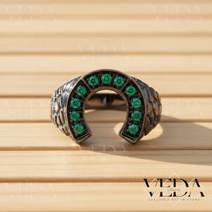 Puede incluir: Un anillo en forma de herradura con un acabado de metal oscuro. El anillo está adornado con numerosas piedras preciosas verdes vibrantes. La banda presenta un diseño texturizado. El anillo está sobre una superficie de madera.