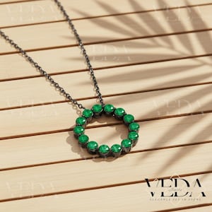 May include: A black chain necklace with a circular pendant. The pendant is a ring of vibrant green gemstones. The necklace is displayed on a light brown wooden surface. The text "VEDA Jewels" and "ELEGANCE SET IN STONE" are visible.