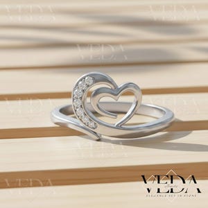 Puede incluir: Un anillo plateado con un diseño de doble corazón. Un corazón está delineado y el otro está lleno de pequeñas piedras preciosas transparentes. El anillo está sobre una superficie de madera. La marca "VEDA Jewels" es visible.
